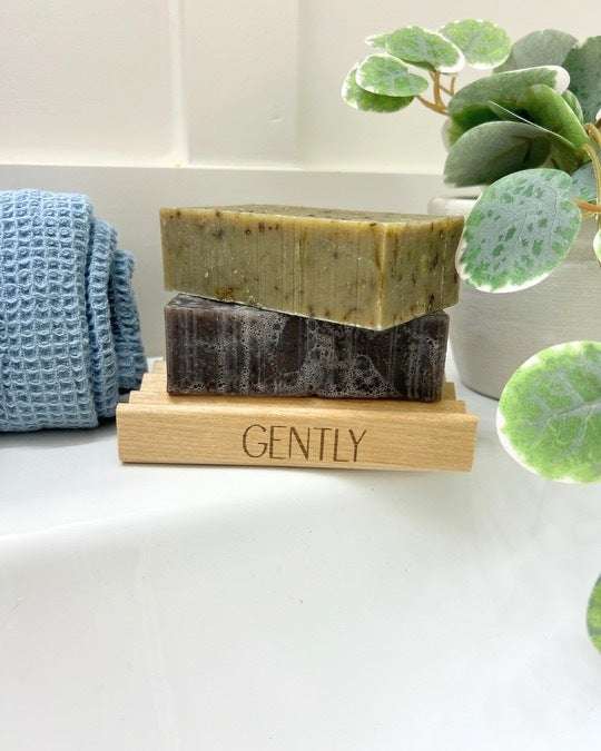 Green and purple bar soap resting on wooden Gently soap tray on a white sink