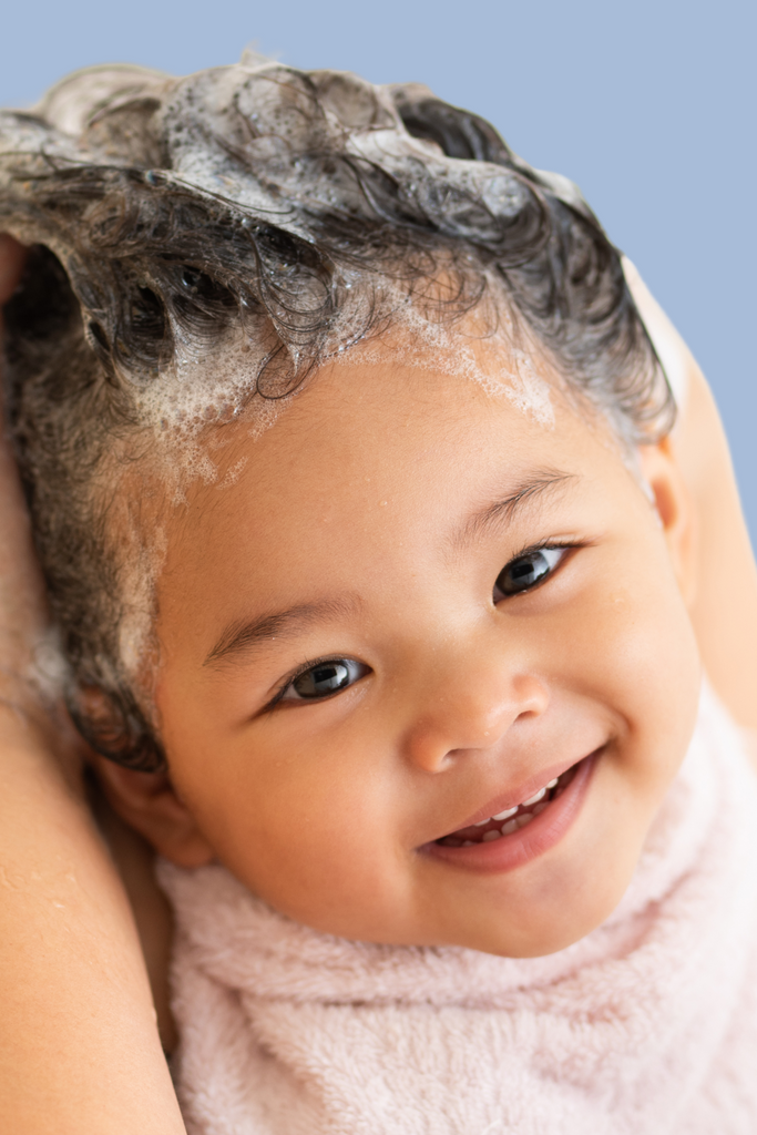 Smiling baby with curly hair covered in foamy shampoo lather during a hair wash, wrapped in a soft towel, against a light blue background