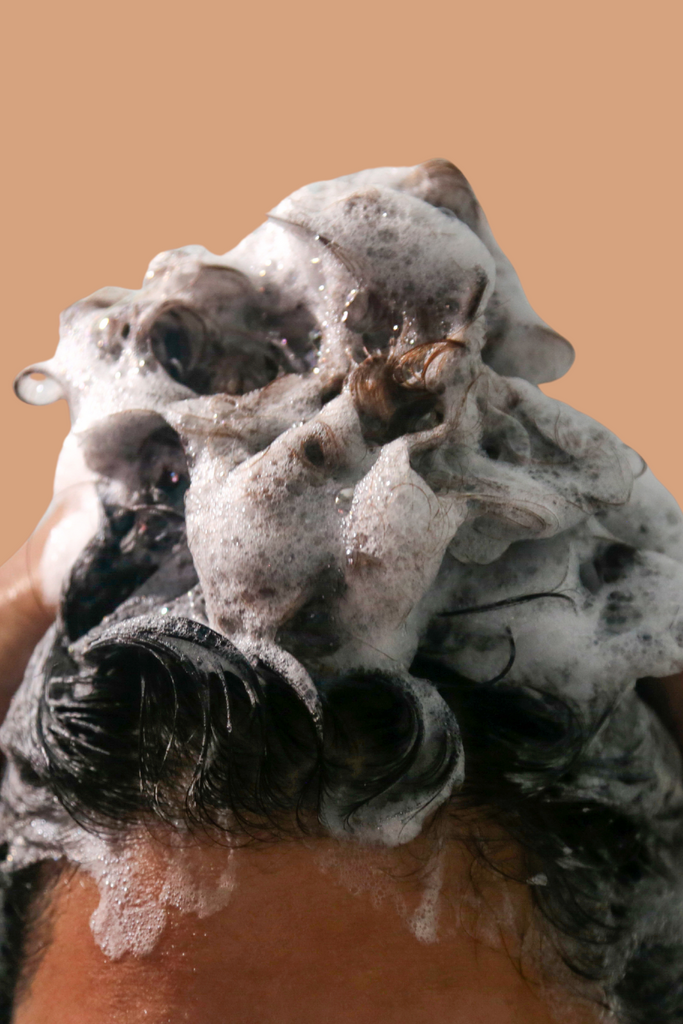 Close-up of brown curly hair covered in thick, foamy shampoo lather against a light orange background
