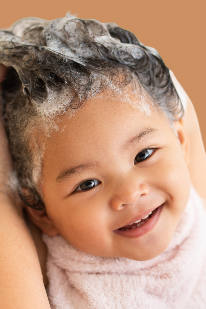 Smiling baby with curly hair covered in foamy shampoo lather during a hair wash, wrapped in a soft towel, against an orange background
