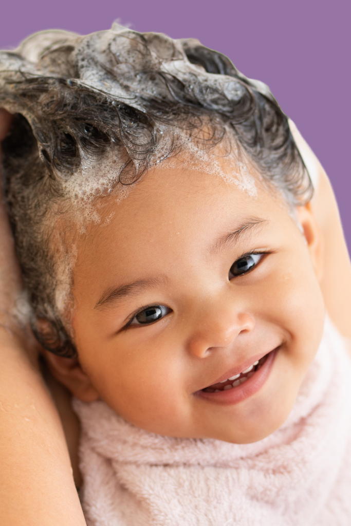 Smiling baby with curly hair covered in foamy shampoo lather during a hair wash, wrapped in a soft towel, against a light purple background